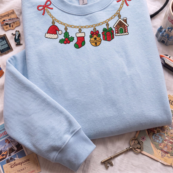 A light blue crewneck sweatshirt laid flat on a white surface, decorated with colorful embroidered Christmas-themed motifs along a gold chain, including a Santa hat, stocking, bell, gift, and gingerbread house. Disney-themed props and a vintage key surround the sweatshirt.