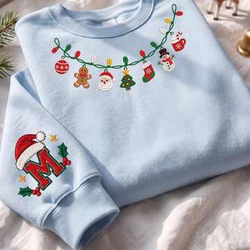 A light blue crewneck sweatshirt laid flat on a white fabric surface, decorated with a Christmas light garland graphic across the chest and a large embroidered letter M with holiday accents on the left sleeve.