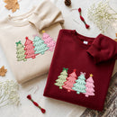 Two folded crewneck sweatshirts with embroidered Christmas trees, one in beige and one in burgundy, styled on a neutral fabric with dried leaves, baby's breath, and embroidery floss.