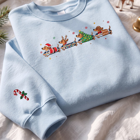 A light blue crewneck sweatshirt laid flat on a white fabric surface. The front has a colorful graphic of four dachshund-shaped Christmas vehicles, and the left sleeve has a small candy cane with holly print.