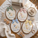 Four personalized oval embroidery hoops featuring embroidered ducks with bows and names, arranged on a white textured blanket with baby-themed props including baby booties, books, wooden blocks, and dried flowers.