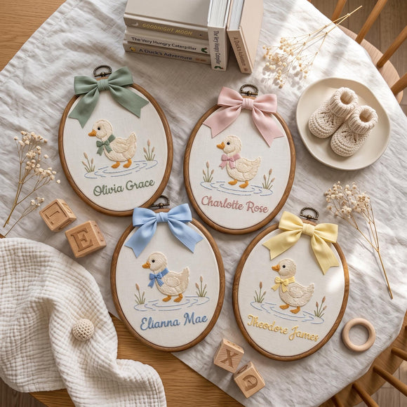 Four personalized oval embroidery hoops featuring embroidered ducks with bows and names, arranged on a white textured blanket with baby-themed props including baby booties, books, wooden blocks, and dried flowers.