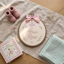 A styled flat-lay featuring a circular embroidery hoop with a personalized fairy-themed design reading 'Aria Rose' in pink script, decorated with a pink bow, floral details, and a fairy illustration. The hoop is surrounded by pink knit baby booties, a pink book titled 'Aria's Sweet Dreams', a mint green muslin cloth, and small dried flowers on a beige linen cloth over a wooden surface.