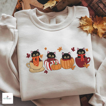 A folded off-white crewneck sweatshirt with a colorful graphic of four black cats sitting in red, yellow, and beige mugs surrounded by autumn leaves. The sweatshirt is styled on a wooden tray with scattered fall leaves, and a 'NUNICAUL' brand logo is visible in the bottom-left corner.