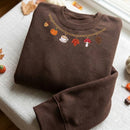 Brown embroidered sweatshirt featuring a gold charm necklace design around the neckline with autumn charms including a pumpkin, acorn, coffee cup, maple leaf, mushroom, and pinecone.