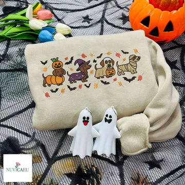 Beige embroidered sweatshirt featuring four Dachshund dogs in Halloween costumes with pumpkin, witch hat, trick-or-treat bucket, and mummy wrap surrounded by flying bats and falling autumn leaves.