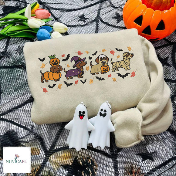 Beige embroidered sweatshirt featuring four Dachshund dogs in Halloween costumes with pumpkin, witch hat, trick-or-treat bucket, and mummy wrap surrounded by flying bats and falling autumn leaves.
