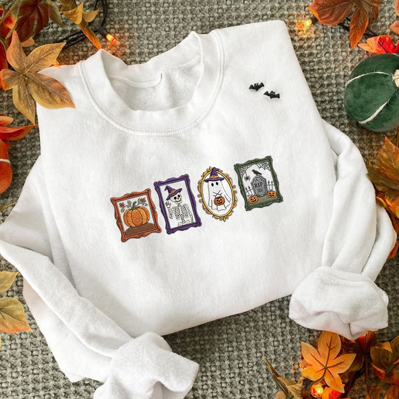 A white long-sleeve sweatshirt laid flat on a textured gray surface, decorated with autumn leaves and small string lights. The front has a horizontal embroidered 'HAPPY HALLOWEEN' design with four character panels: a pumpkin, a witch in a purple frame, a ghost in a gold frame, and a haunted house in a green frame.