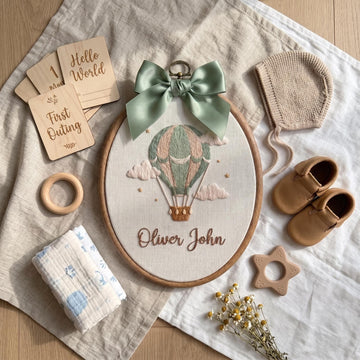 A styled flat-lay of a personalized embroidery hoop featuring a hot air balloon design with the text 'Oliver John' and a sage green ribbon bow. The hoop is surrounded by baby-themed wooden and fabric accessories, including a knit bonnet, moccasins, a teething ring, a muslin cloth, and wooden name cards on a neutral linen background.
