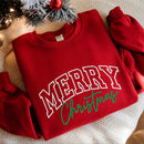 A folded red crewneck sweatshirt with 'MERRY Christmas' printed on the front, styled on a textured cream blanket with a decorated Christmas tree branch in the corner.