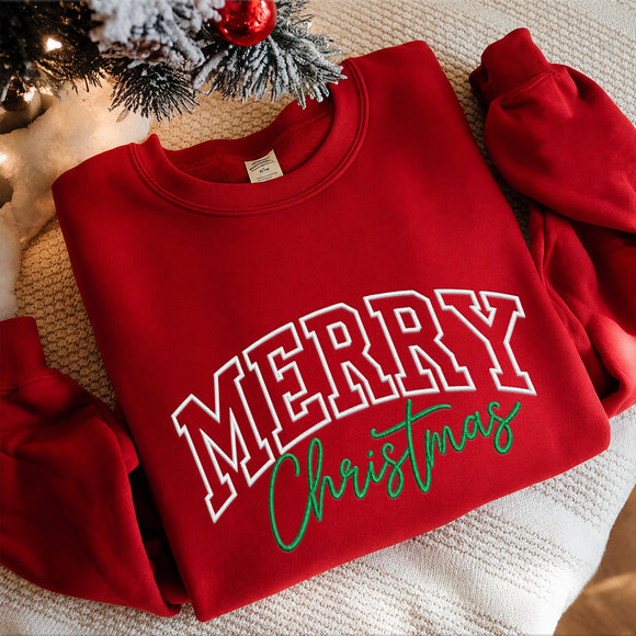 A folded red crewneck sweatshirt with 'MERRY Christmas' printed on the front, styled on a textured cream blanket with a decorated Christmas tree branch in the corner.