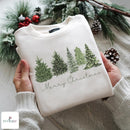 A folded cream-colored crewneck sweatshirt with a green Christmas tree graphic and 'Merry Christmas' text, held by hands wearing a matching cream knit sweater. The background features festive green pine branches, snow-dusted pinecones, and red berries, with a 'NUVICAEMU' brand logo in the bottom-left corner.