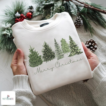 A folded cream-colored crewneck sweatshirt with a green Christmas tree graphic and 'Merry Christmas' text, held by hands wearing a matching cream knit sweater. The background features festive green pine branches, snow-dusted pinecones, and red berries, with a 'NUVICAEMU' brand logo in the bottom-left corner.