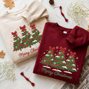 Two folded crewneck sweatshirts with embroidered Christmas tree designs and 'Merry Christmas' text, styled on a neutral fabric background with autumn leaves, baby's breath, and embroidery thread.