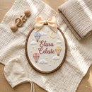 Personalized wooden embroidery hoop with a pink bow, featuring an embroidered rainbow, clouds, hearts, and the name 'Elara Fern' on a white fabric background, styled in a nursery setting.