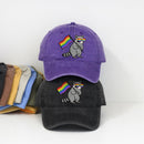 Two baseball caps displayed on a white background, one purple and one black, both featuring an embroidered raccoon wearing sunglasses and holding a rainbow flag. A stack of other colored caps is partially visible on the left.