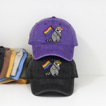 Two baseball caps displayed on a white background, one purple and one black, both featuring an embroidered raccoon wearing sunglasses and holding a rainbow flag. A stack of other colored caps is partially visible on the left.
