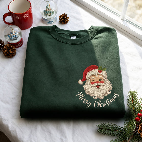 A dark green crewneck sweatshirt with an embroidered Santa Claus face and 'Merry Christmas' text, folded on a white fabric surface with holiday props including a red mug, snow globes, pinecones, and evergreen branches.