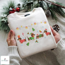 A folded cream-colored crewneck sweatshirt with a festive Christmas dachshund graphic, held by two hands wearing a matching knit sweater. The background is decorated with pine branches, pinecones, and red berries, creating a cozy holiday atmosphere.