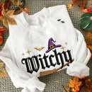 A white long-sleeve sweatshirt with a Halloween-themed graphic print featuring the word 'Witchy' in black gothic lettering, a purple witch hat, gold stars, bats, and a broomstick. The sweatshirt is styled in a flat-lay with autumn leaves and fairy lights on a gray textured background.