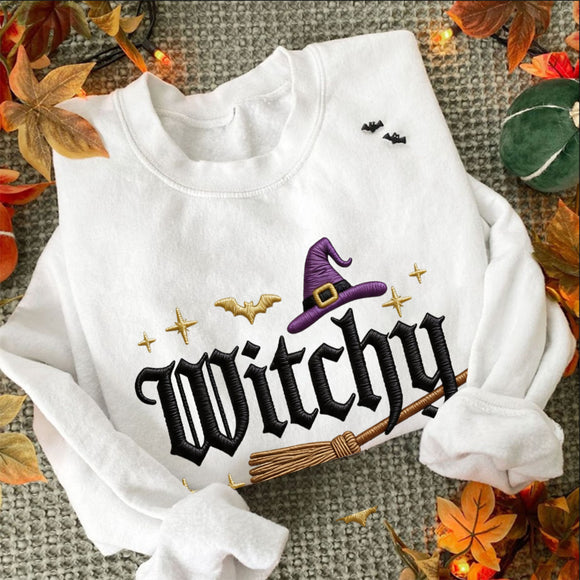 A white long-sleeve sweatshirt with a Halloween-themed graphic print featuring the word 'Witchy' in black gothic lettering, a purple witch hat, gold stars, bats, and a broomstick. The sweatshirt is styled in a flat-lay with autumn leaves and fairy lights on a gray textured background.