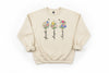 Beige sweatshirt with floral designs and text 'faith', 'hope', 'love' on a wooden surface.