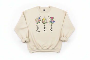 Beige sweatshirt with floral designs and text 'faith', 'hope', 'love' on a wooden surface.