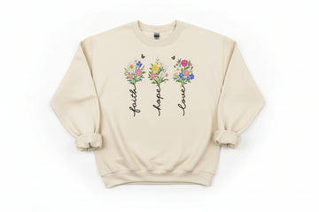 Beige sweatshirt with floral designs and text 'faith', 'hope', 'love' on a wooden surface.