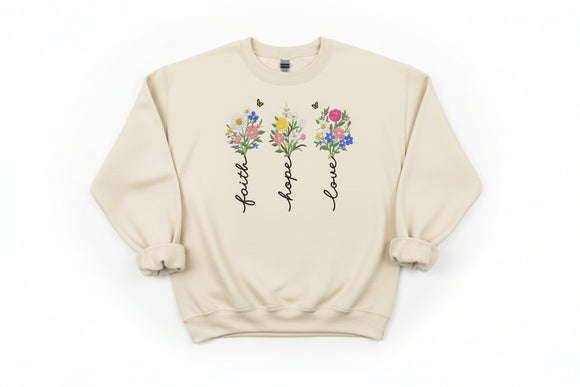 Beige sweatshirt with floral designs and text 'faith', 'hope', 'love' on a wooden surface.