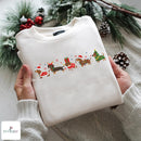 A folded cream-colored crewneck sweatshirt with a colorful Christmas dachshund graphic, held by two hands wearing a matching cream knit sweater. The background is decorated with pine branches, snow-dusted greenery, and red berries, and a 'NUVIAEU' brand logo is visible in the bottom-left corner.