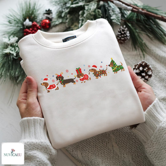 A folded cream-colored crewneck sweatshirt with a colorful Christmas dachshund graphic, held by two hands wearing a matching cream knit sweater. The background is decorated with pine branches, snow-dusted greenery, and red berries, and a 'NUVIAEU' brand logo is visible in the bottom-left corner.
