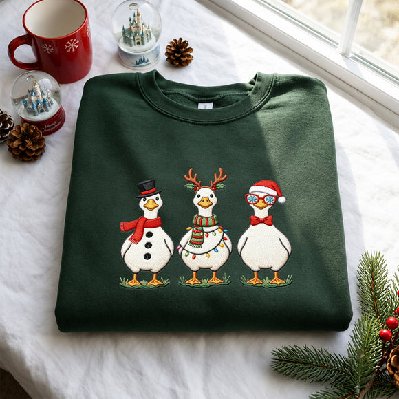 A dark green crewneck sweatshirt folded on a white fabric surface, featuring a festive graphic of three cartoon ducks dressed as snowmen. The scene is styled with holiday props including a red mug, snow globes, pinecones, and evergreen branches near a window.