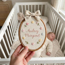 A hand holds an oval embroidery hoop featuring personalized embroidered text reading 'Audrey Margaret' with small pastel flowers and green leaves on a cream fabric background, tied with a bow at the top. A sleeping newborn baby in a cream outfit rests in a white crib in the softly lit background.