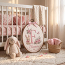 A styled nursery scene with a white crib, pink floral bedding, a cream cable-knit throw, a plush bunny, a woven basket with rolled pink blankets, and a large oval embroidered wooden sign leaning in front. The sign features a pink letter F, floral details, a bunny illustration, and the name Eleanor with a date.