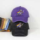 Two baseball caps stacked on a white background, with a purple cap on top and a black cap underneath. Both caps feature an embroidered raccoon wearing a rainbow cape and holding a sword, with the text 'we pride at dawn' below.