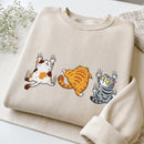 A folded cream-colored sweatshirt featuring a graphic print of three cartoon cats in different poses, displayed on a light surface with small white flowers and a textured fabric in the background.