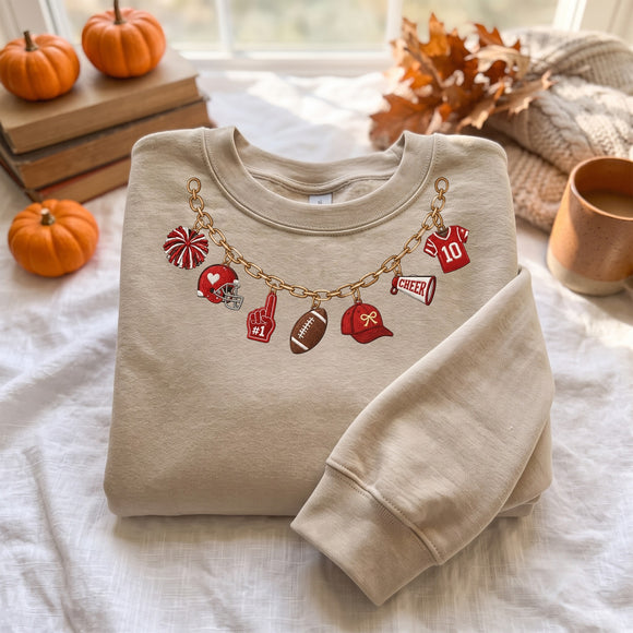 A beige crewneck sweatshirt with a gold chain and red sports-themed charms, styled on a white fabric surface with autumn props like pumpkins, books, and a mug.
