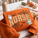 An orange sweatshirt with a Halloween-themed 'NURSE' graphic, featuring cartoon ghosts, bats, spiderwebs, and checkered lettering, styled on a table with autumn props.