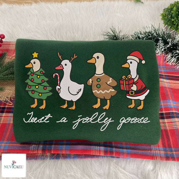 A square dark green embroidered pillow featuring four festive goose characters in holiday outfits and the phrase 'Just a jolly goose' in white script. The pillow is styled on a red plaid blanket over a white faux fur surface, with green pine branches in the background.
