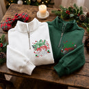 Two Christmas-themed quarter-zip sweatshirts are displayed on a rustic wooden table. One is white and the other is forest green, both featuring a 'Joyful Merry 2023' graphic with red and green holiday lettering and holly accents.