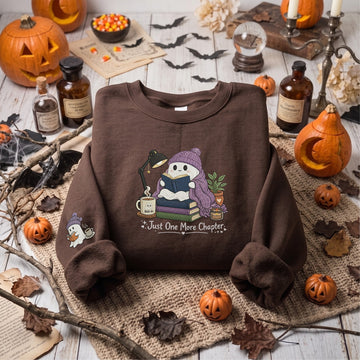 A brown crewneck sweatshirt with a colorful Halloween graphic featuring cats, pumpkins, a witch, and a crescent moon, displayed in a styled flat-lay setting with pumpkins, candles, and autumn leaves.