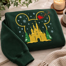 A dark green crewneck sweatshirt featuring a large embroidered Mickey Mouse silhouette with a Christmas theme, including a gold castle, snow trees, and a red sleigh. The sweatshirt is styled in a cozy flat-lay setting with a candle, book, and knit blanket.