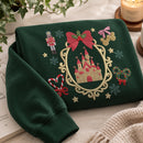 A folded dark green sweatshirt with a large embroidered Christmas-themed design featuring a castle in an ornate frame, red bows, snowflakes, a nutcracker, candy canes, and gold stars, styled on a light fabric surface with a candle, book, and plant nearby.