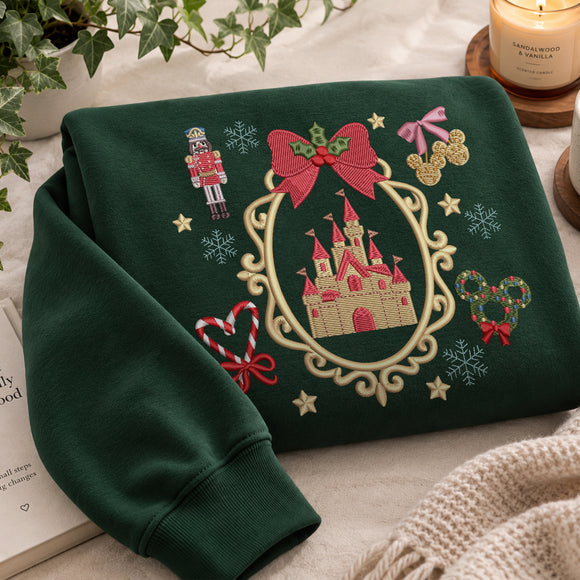 A folded dark green sweatshirt with a large embroidered Christmas-themed design featuring a castle in an ornate frame, red bows, snowflakes, a nutcracker, candy canes, and gold stars, styled on a light fabric surface with a candle, book, and plant nearby.