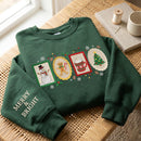 A dark green crewneck sweatshirt with festive Christmas graphics, laid flat on a woven wicker table. The sweatshirt features four embroidered holiday-themed panels (a snowman, a gingerbread man, a mug of hot cocoa, and a Christmas tree) across the chest, 'MERRY & BRIGHT' embroidered in gold on the left sleeve, and is styled next to a spool of thread and a ceramic mug.