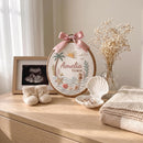 Personalized circular embroidery hoop art featuring the name 'Amelia Grace' in a beach-themed design with a pink ribbon bow, styled on a wooden dresser alongside a framed ultrasound photo, knit booties, a shell-shaped dish, dried flowers, and a folded blanket.