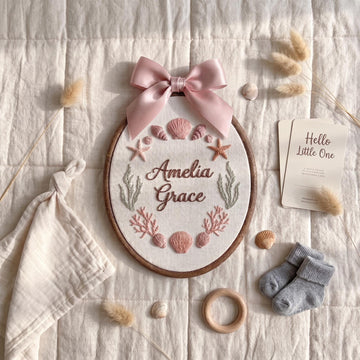 Personalized oval embroidery hoop wall art with a pink satin bow, featuring the names 'Amelia Grace' and an ocean-themed design with shells, starfish, coral, and seagrass. The hoop is styled in a flat lay with baby socks, a wooden ring, dried grass, a muslin cloth, and a card reading 'Hello Little One' on a cream quilted fabric background.