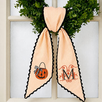 Personalized peach and white striped fabric bow with black scalloped edges, featuring a pumpkin design on one side and a monogrammed letter 'M' on the other, displayed on a green wreath in front of a window.