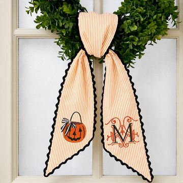 Personalized peach and white striped fabric bow with black scalloped edges, featuring a pumpkin design on one side and a monogrammed letter 'M' on the other, displayed on a green wreath in front of a window.