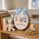 Personalized wooden embroidery hoop sign with a light blue satin bow, featuring embroidered baby clothes, a teddy bear, and the name 'Noah Thomas' on a wooden shelf next to a knitted teddy bear and books.
